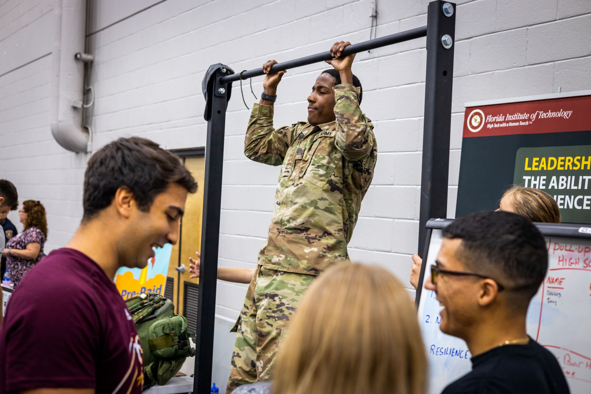 A military personnel performing pull-ups at a Florida Tech event. Other attendees are gathered around, watching and engaging in conversation. A sign in the background reads 'Florida Institute of Technology' and highlights leadership and resilience
